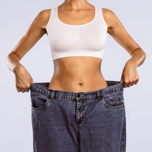 Closeup cropped view of skinny woman showing flat stomach pulling oversized big blue pants jeans. Slim body low fat. Healthy size athletic posing girl isolated over blue color studio background